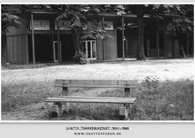 Das Foto zeigt den im April 1944 errichteten hölzernen Kinderpavillon kurz nach der Befreiung im Jahr 1945. Die Glasfronten sind nur noch teilweise vorhanden.  Tafeln mit Tiermotiven, die den Bau schmücken sollten, sind jedoch noch erhalten (an der der linken Außenfront  u. a. Hasen und Bären). Die Bilder  stammen von dem niederländischen Künstler Jo Spier. Die Bank im Vordergrund wurde ebenfalls im Zuge der Neugestaltung des Parks  im Jahr 1944 aufgestellt.  © Památník Terezín A 1902, Fotograf Jiří Lauscher