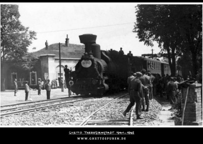 Ein Personenzug, versehen mit Girlanden und der Aufschrift „1.6.1943“, kommt in Theresienstadt an. Das Foto hält den Moment der Einweihung der Bahnstrecke fest. Im Hintergrund die Wälle der Festung am südlichen Stadtrand, wo sich ehemals das Bauschowitzer Tor befand. © Yad Vashem